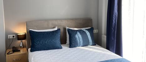 A hotel room with a bed, bedside tables, a lamp, an air conditioning unit, and a window with curtains.