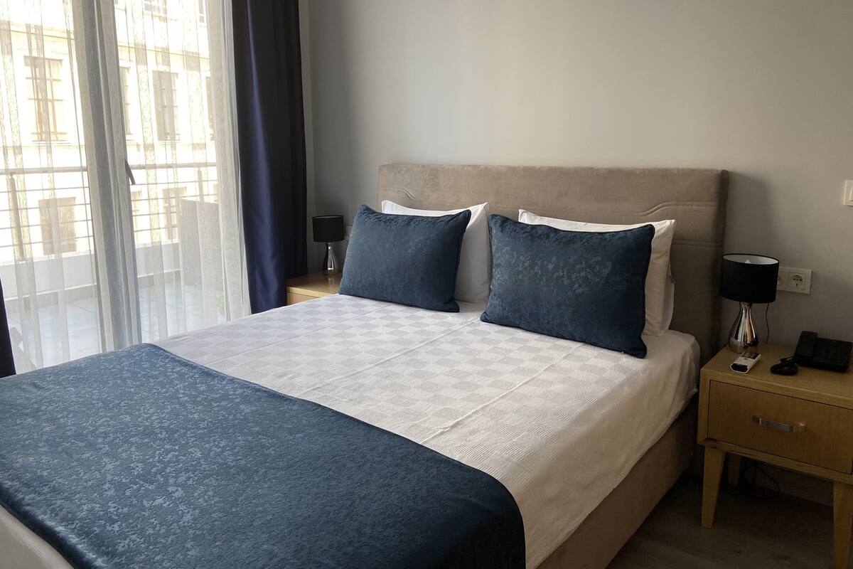 A neatly made bed with a patterned bedspread, two pillows, a bedside table with a lamp, and a window with curtains.