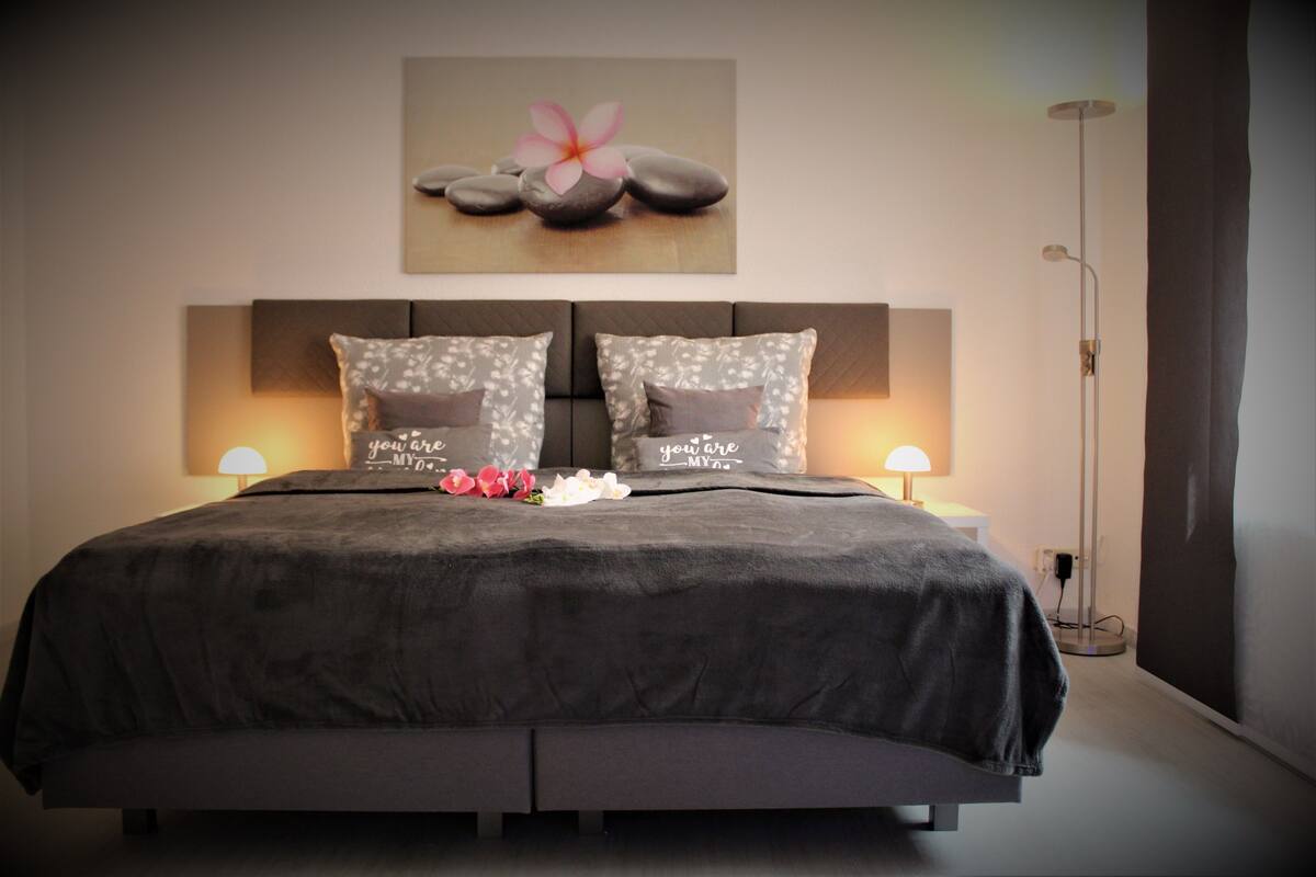 A neatly made bed with a dark bedspread and two pillows, a wall-mounted artwork of stones and a flower, and two bedside lamps.