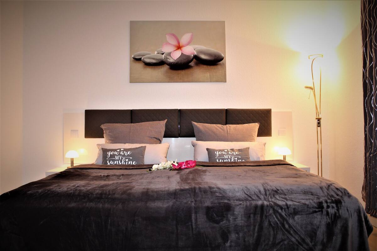A neatly made bed with a dark bedspread, two pillows with a message, a wall art of stones and a flower, and a bedside lamp.