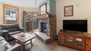 A living room with a stone fireplace, leather sofas, a television, and a view of a deck with a ski lift. - River Run Condo with Slope Views, Short Walk to Lifts & Close to Gondola (Keystone)