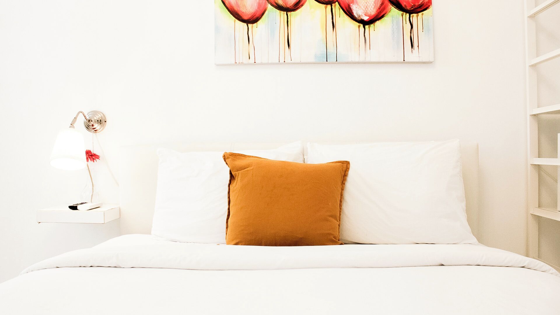 Un lit avec une literie blanche et un oreiller de couleur moutarde, une table de chevet blanche avec une lampe, et un tableau accroché au mur au-dessus du lit.