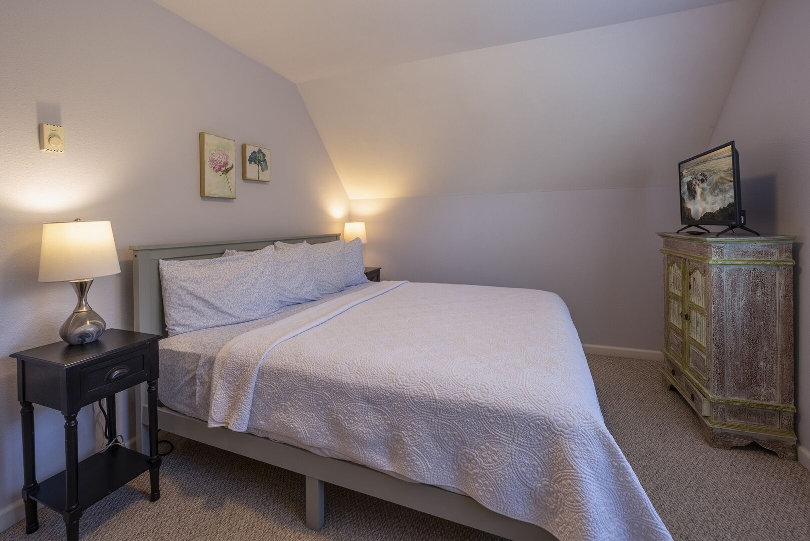 A bedroom with a bed, bedside lamps, a nightstand, a dresser, and wall art.