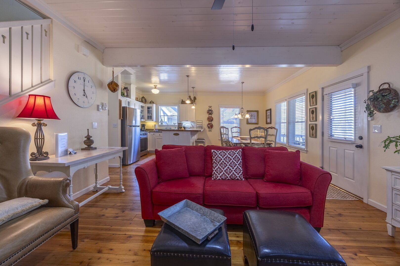 Un salon chaleureux avec un canapé rouge, une table basse en bois et un fauteuil en cuir. On aperçoit, au fond, une cuisine avec des meubles blancs et un coin repas comprenant une table et des chaises.