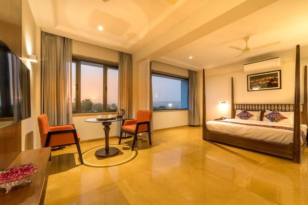 Habitación de hotel con cama con dosel, una mesa de comedor con sillas, una pequeña mesa redonda y vista a la ciudad al anochecer.