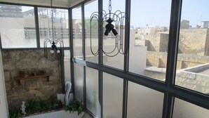 A high-ceilinged room with large windows, a wooden shelf with boxes, a small plant, and a hanging light fixture. - Tal-Kapilluna Zebbug Guest House (Zebbug)