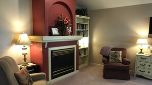 Ein Wohnzimmer mit Kamin, Bücherregal, zwei Sesseln und zwei Nachttischen mit Lampen.