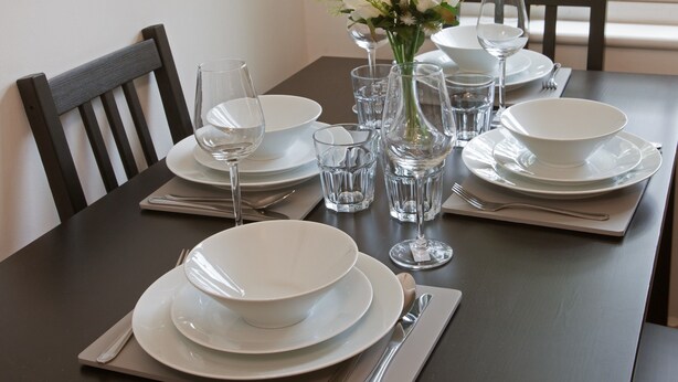 A dining table set with white plates, bowls, glasses, and cutlery, with a vase of flowers in the center. - Victoria's place (Chelmsford)