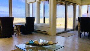 Ein Zimmer mit Glastisch, der dekorative Gegenstände zeigt, Blick auf den Strand durch große Fenster und eine Sitzecke mit Stühlen.