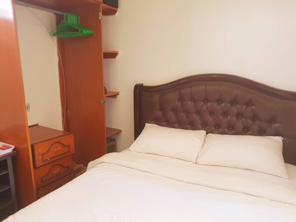 A neatly made bed with a tufted headboard, a wooden nightstand, and a wardrobe with a mirror and hanging clothes.