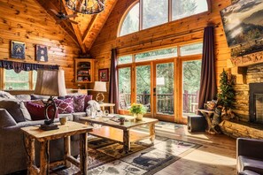 A cozy log cabin interior with a fireplace, large windows, and a wooden ceiling.