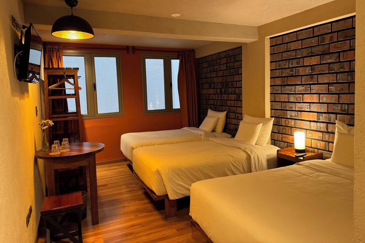 A hotel room with two beds, a wooden desk, a hanging coat rack, and a brick-patterned wall.