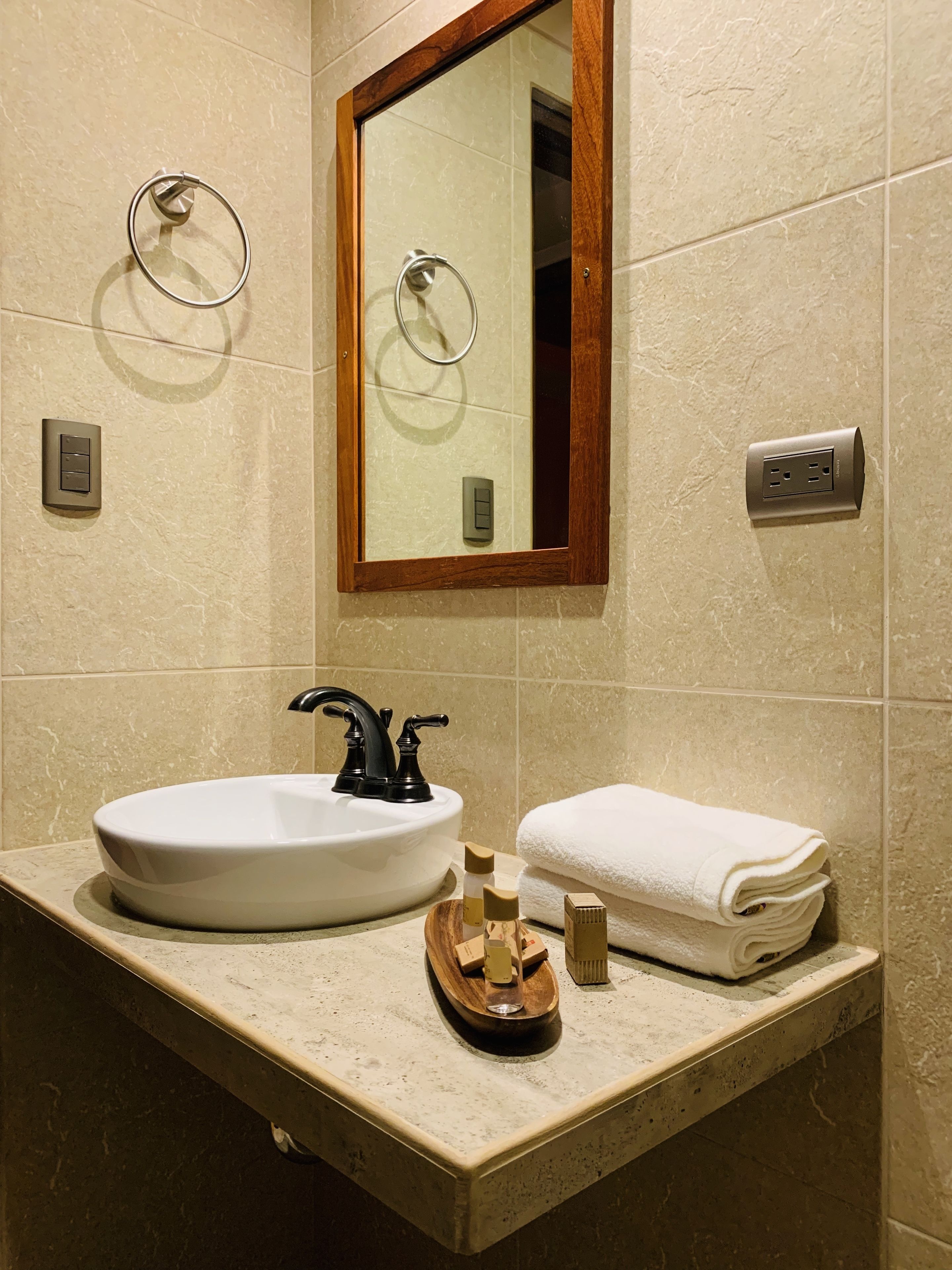 Un baño con un lavabo blanco, grifo negro y una bandeja de madera con artículos de aseo. Hay un espejo sobre el lavabo y una toalla en la encimera.
