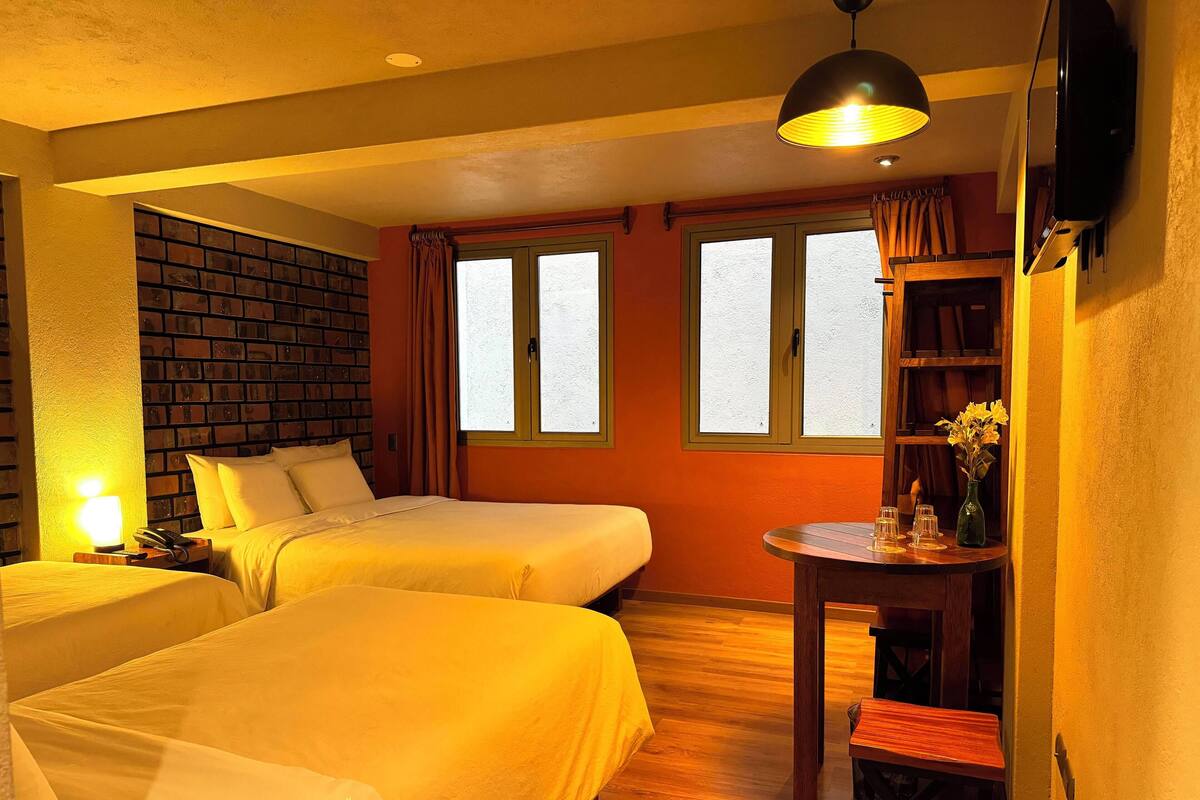 A hotel room with two beds, a wooden floor, a brick accent wall, a desk with a chair, and a TV mounted on the wall.