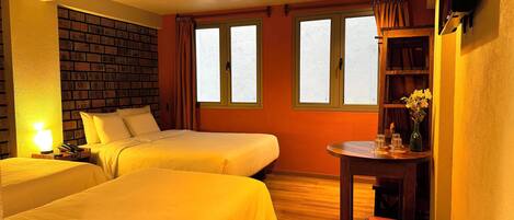 A hotel room with two beds, a wooden floor, a brick accent wall, a desk with a chair, and a TV mounted on the wall.