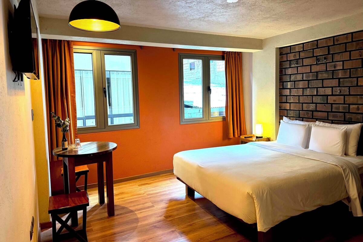 A hotel room with a bed, a desk with a chair, two windows with curtains, a brick-patterned headboard, and a hanging light fixture.