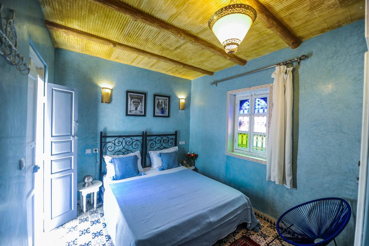 A bedroom with a bed, a window with curtains, a wall-mounted light, and a door.