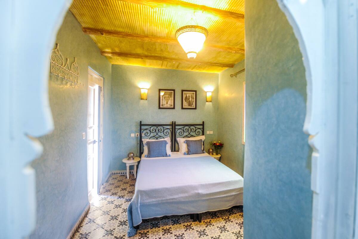 A bedroom with a bed, two bedside tables, a nightstand, a chandelier, and framed pictures on the wall.