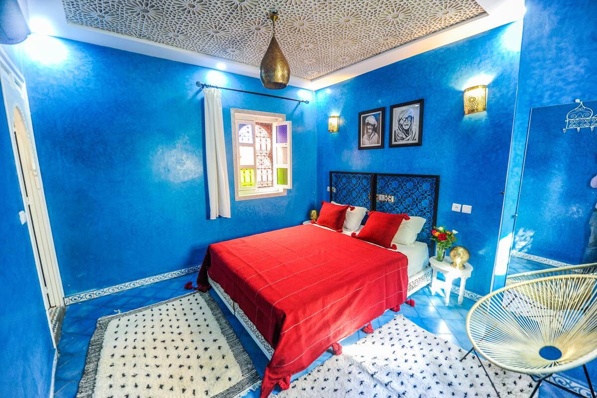A bedroom with a blue wall, a red bedspread, a white chair, and a decorative ceiling.