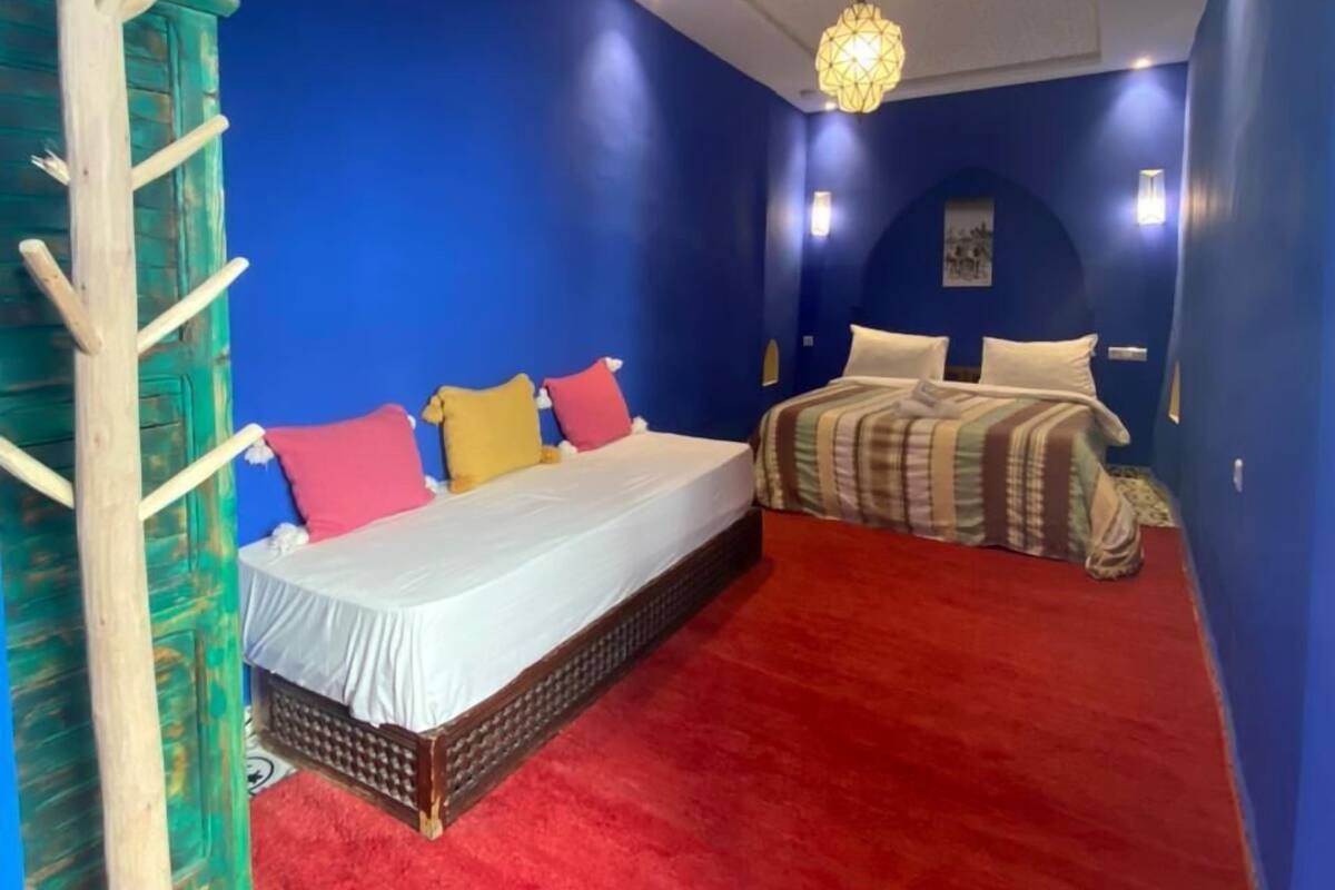 A bedroom with a red carpet, a bed with white bedding and colorful pillows, a wooden ladder, and a decorative wall hanging.