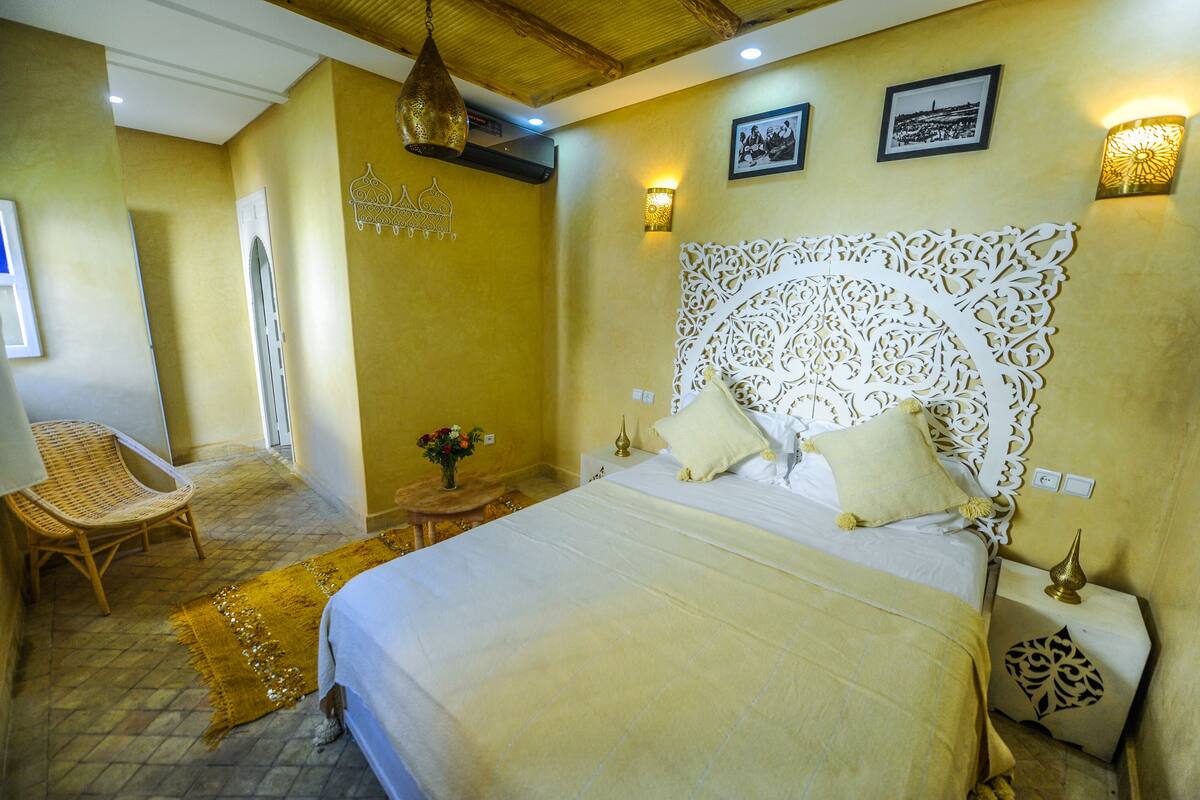 A bedroom with a large bed, a decorative headboard, a wicker chair, a small table with a vase of flowers, and wall-mounted lights.