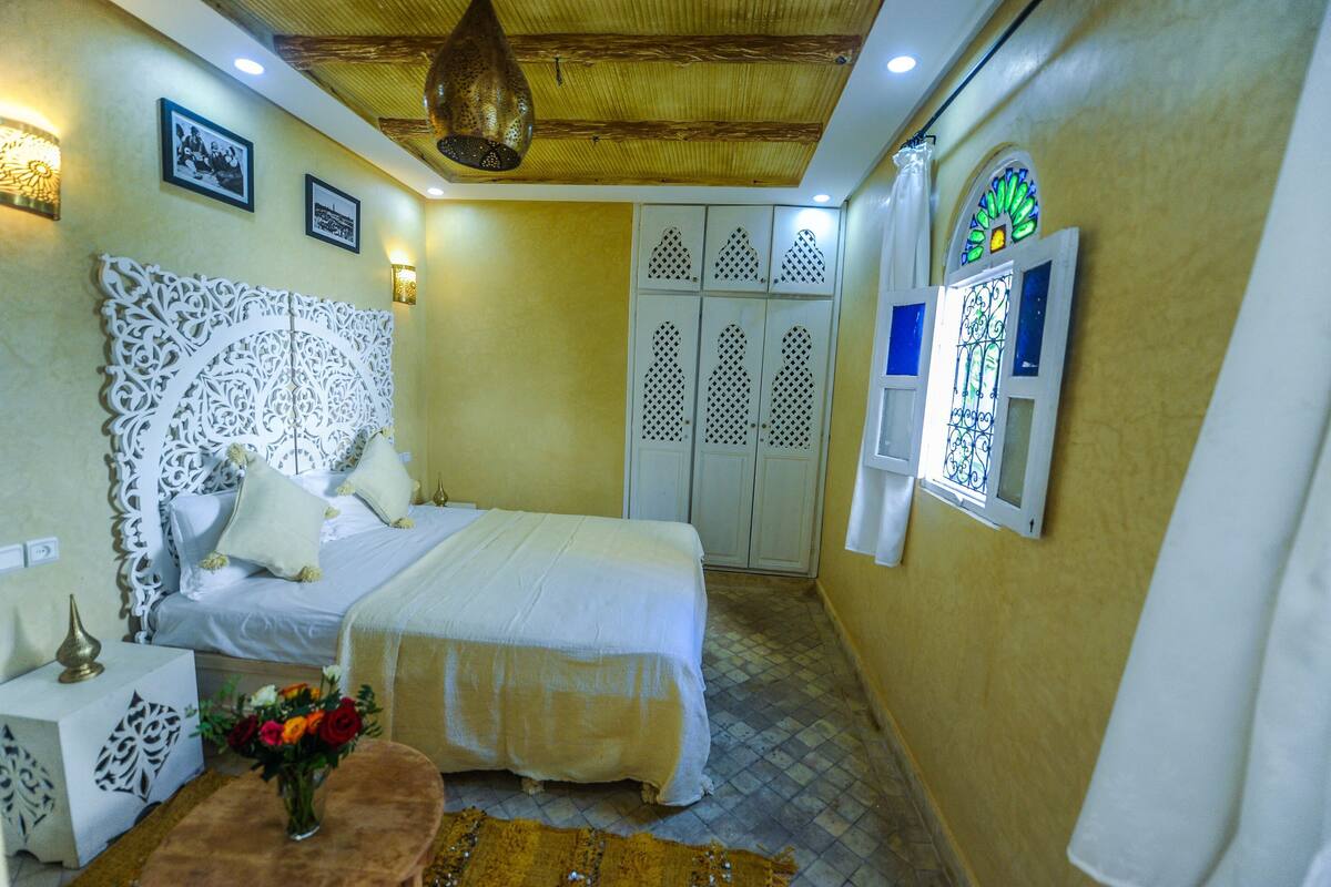 A bedroom with a large ornate headboard, a bed with white linens, a nightstand with a vase of flowers, and a wall-mounted TV.