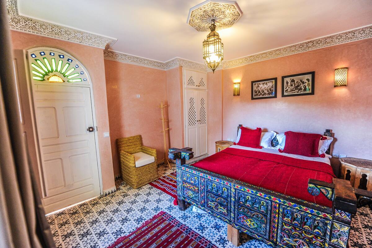 A traditional room with a bed, a chair, a door, and a wall-mounted light fixture.