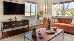 Un salon moderne doté d’un téléviseur à écran plat, d’un canapé, d’une table basse agrémentée d’objets décoratifs et d’une grande fenêtre offrant une vue sur la verdure et les collines.