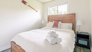 Een net opgemaakt bed met een houten hoofdeinde, witte beddengoed en opgerolde handdoeken aan het voeteneinde. Er staat een nachtkastje met een lamp naast het bed.