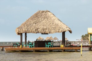 A thatched-roof structure with a wooden deck, chairs, and tables, overlooking the sea.