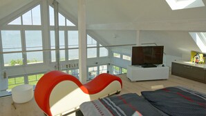 Una stanza con soffitto alto e una grande finestra che si affaccia su un lago, un letto, una televisione e un angolo cottura.