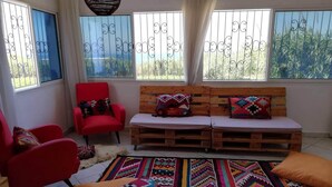 Ein Zimmer mit einer Holzbank, roten Stühlen, bunten Teppichen und Blick durch vergitterte Fenster.