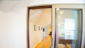 Een gang met houten vloer, een glazen deur en uitzicht op een kamer met raam en prullenbak.
