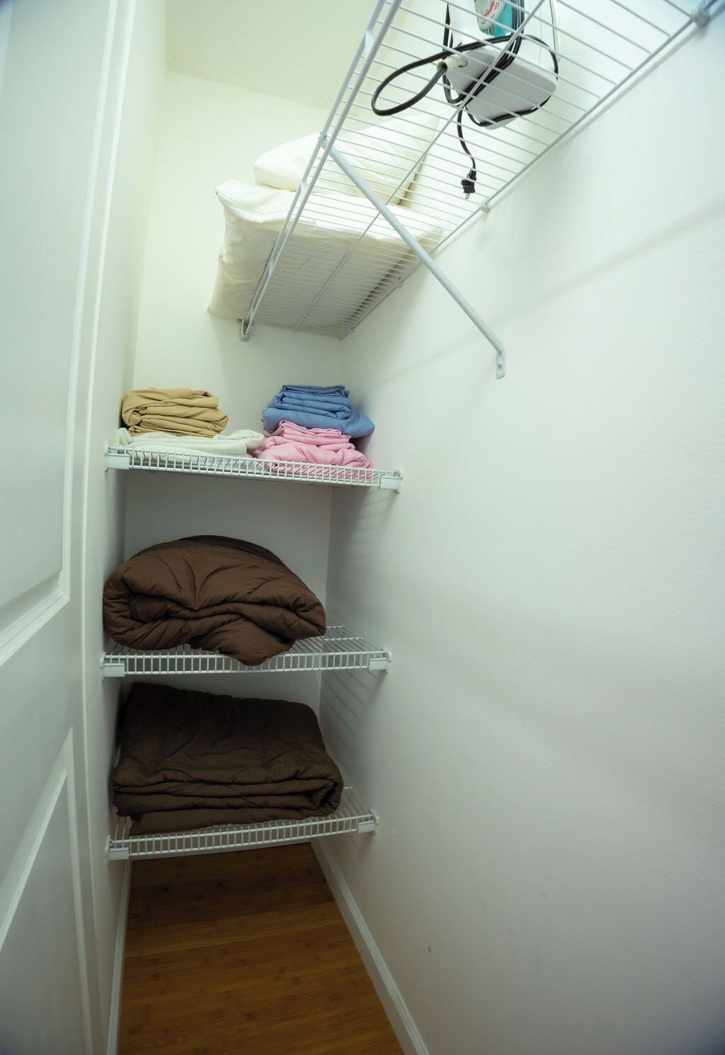 A narrow hallway with white walls, a wooden floor, and a metal shelf unit with folded blankets.
