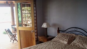Une chambre à coucher comprenant un lit, une table de chevet, une lampe et une porte en bois donnant sur un espace extérieur aménagé avec des chaises.