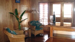 Une pièce aux murs en bois, avec une grande plante, deux fauteuils en osier, une table, une lampe et une bouteille posée sur un comptoir.