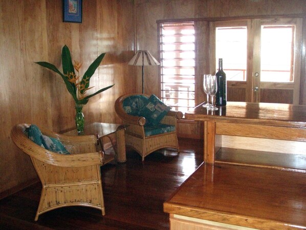 A room with wooden walls, a large plant, two wicker chairs, a table, a lamp, and a bottle on a counter.