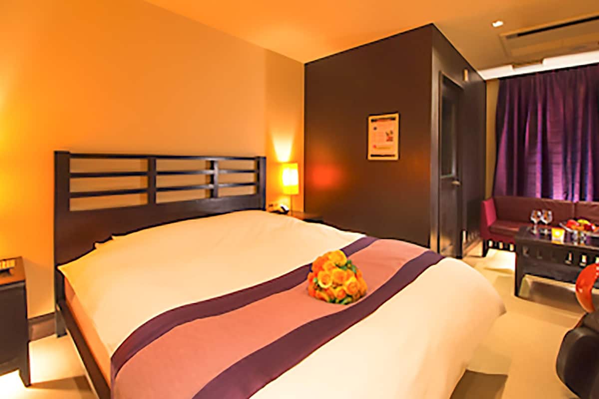 A hotel room with a large bed, a nightstand with a lamp, a seating area with a couch and a table, and a door leading to another room.