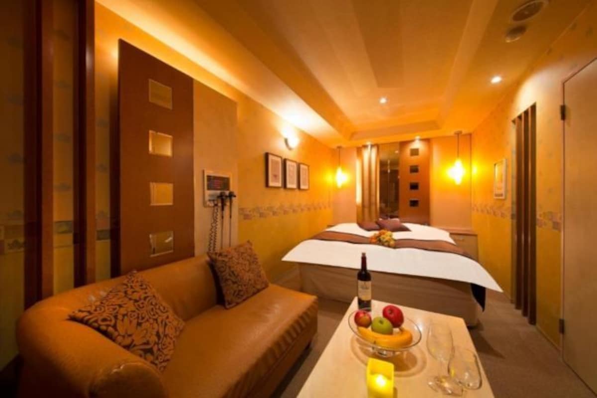 A hotel room with a bed, a sofa, a table with fruit and a bottle, and a bathroom with a shower and toilet.