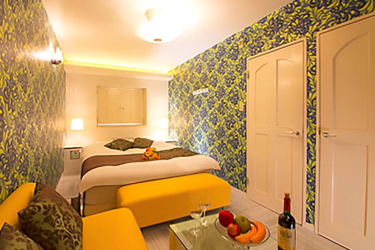 A hotel room with a bed, a sofa, a glass coffee table with fruit and a bottle, and a door.