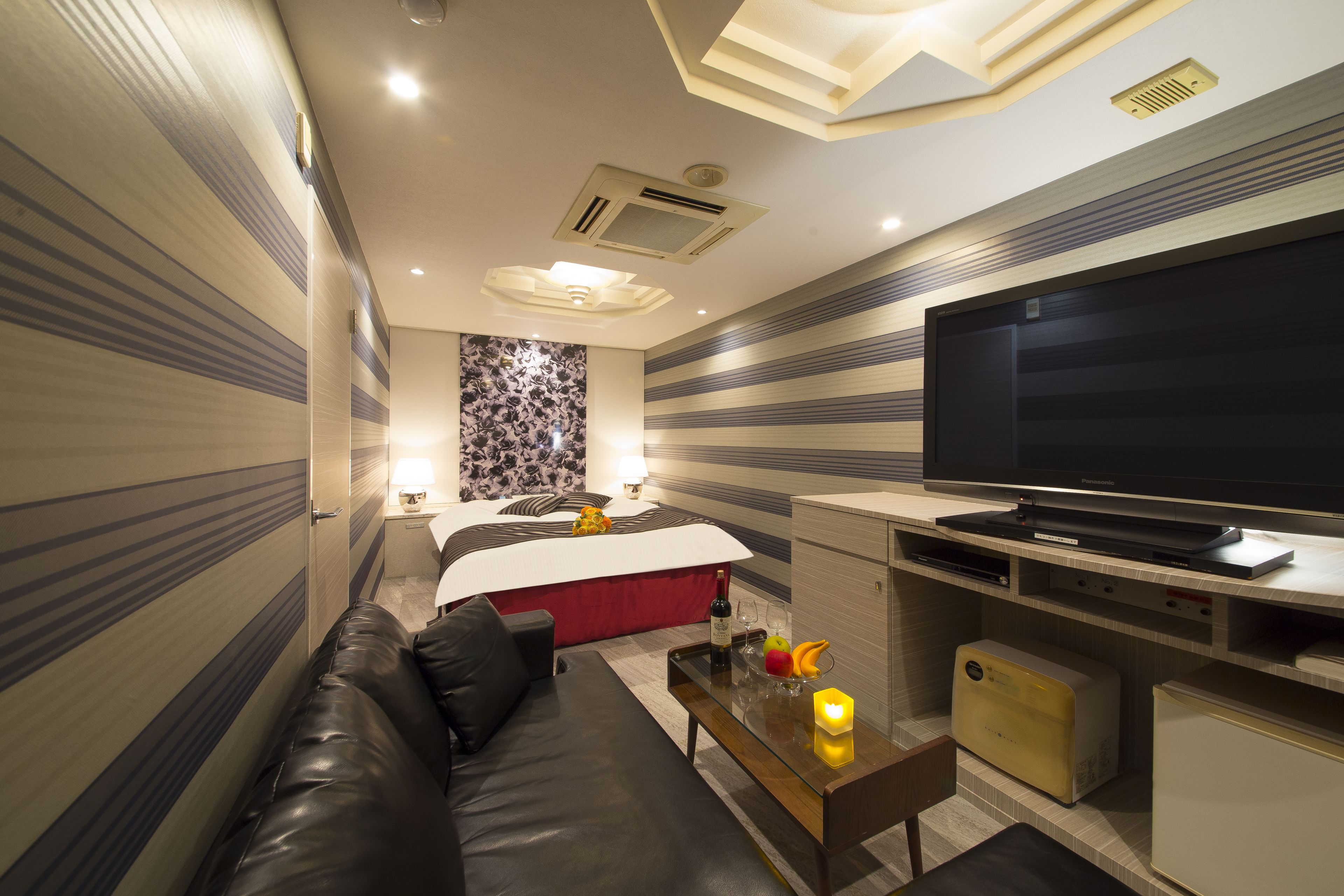 A modern hotel room with a large bed, a flat-screen TV, a leather sofa, a small table with a candle, and a microwave.