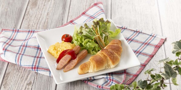 A square plate with scrambled eggs, sausages, a croissant, a small salad, and a cherry tomato on a plaid cloth.