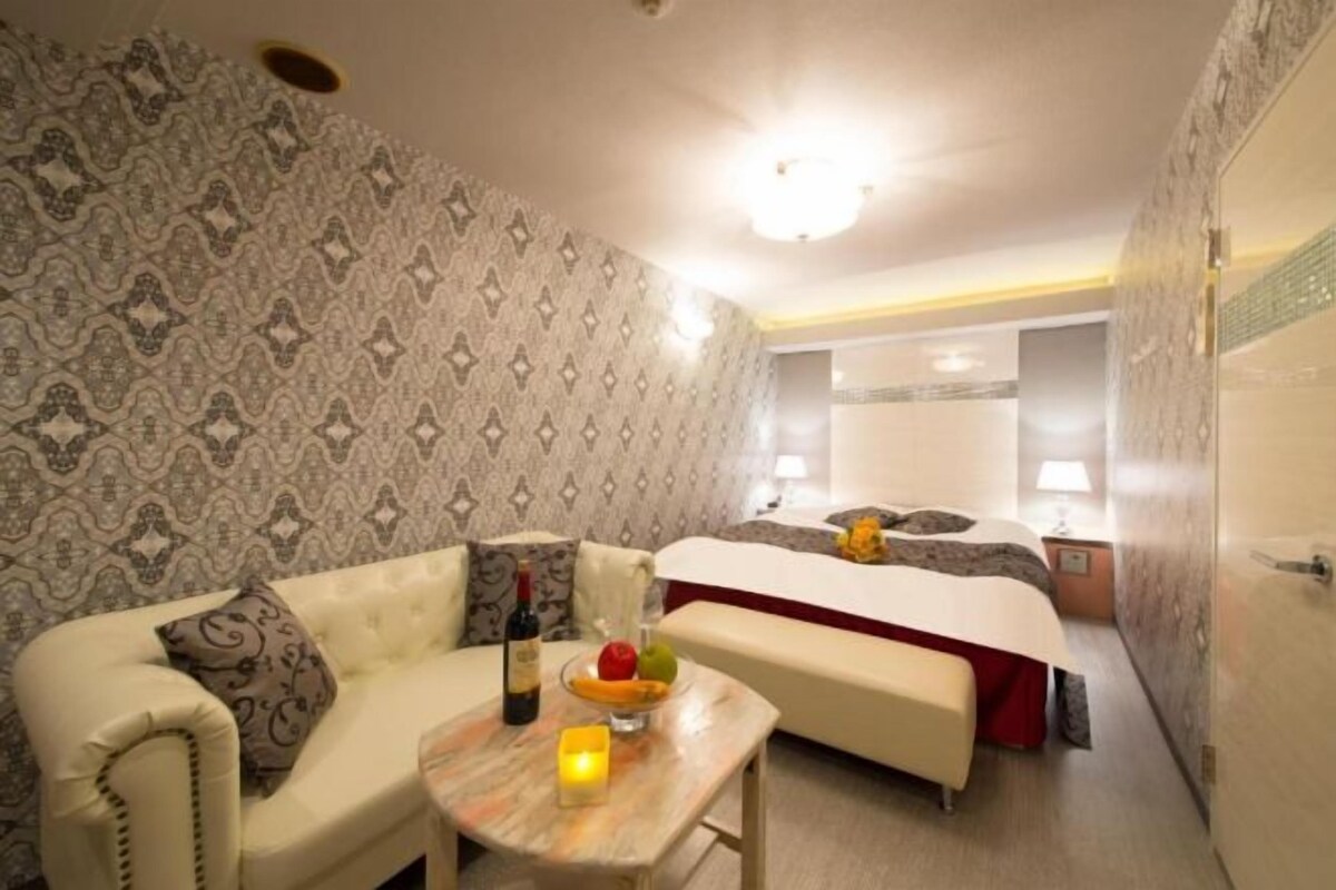 A hotel room with a patterned wallpaper, a sofa, a small table with a candle, a fruit bowl, and a bed with a red bedspread.