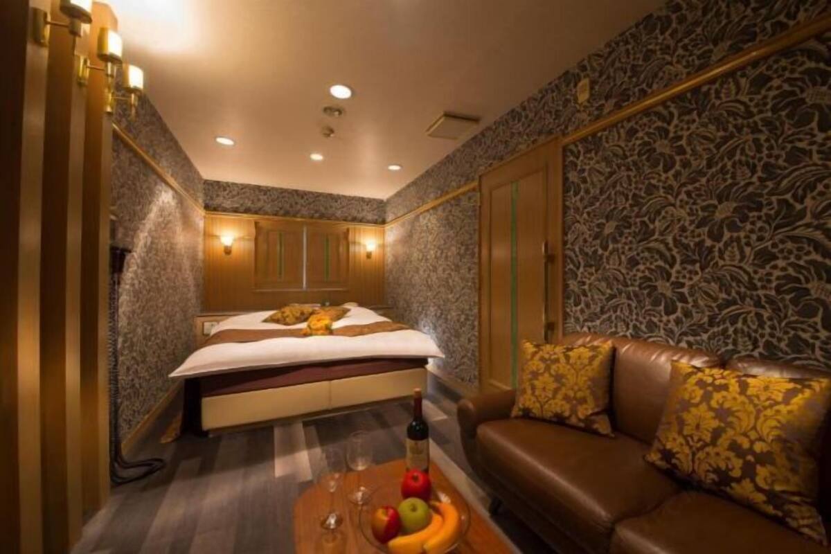 A hotel room with a bed, a sofa, a coffee table with fruits and a bottle, and patterned wallpaper.