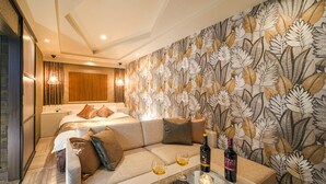 A hotel room with a bed, a sofa, a glass table with a bottle and glasses, and a wall with a leaf pattern.