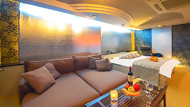 A modern hotel room with a large sofa, a bed, a dining table with fruit and wine, and a view of the city.
