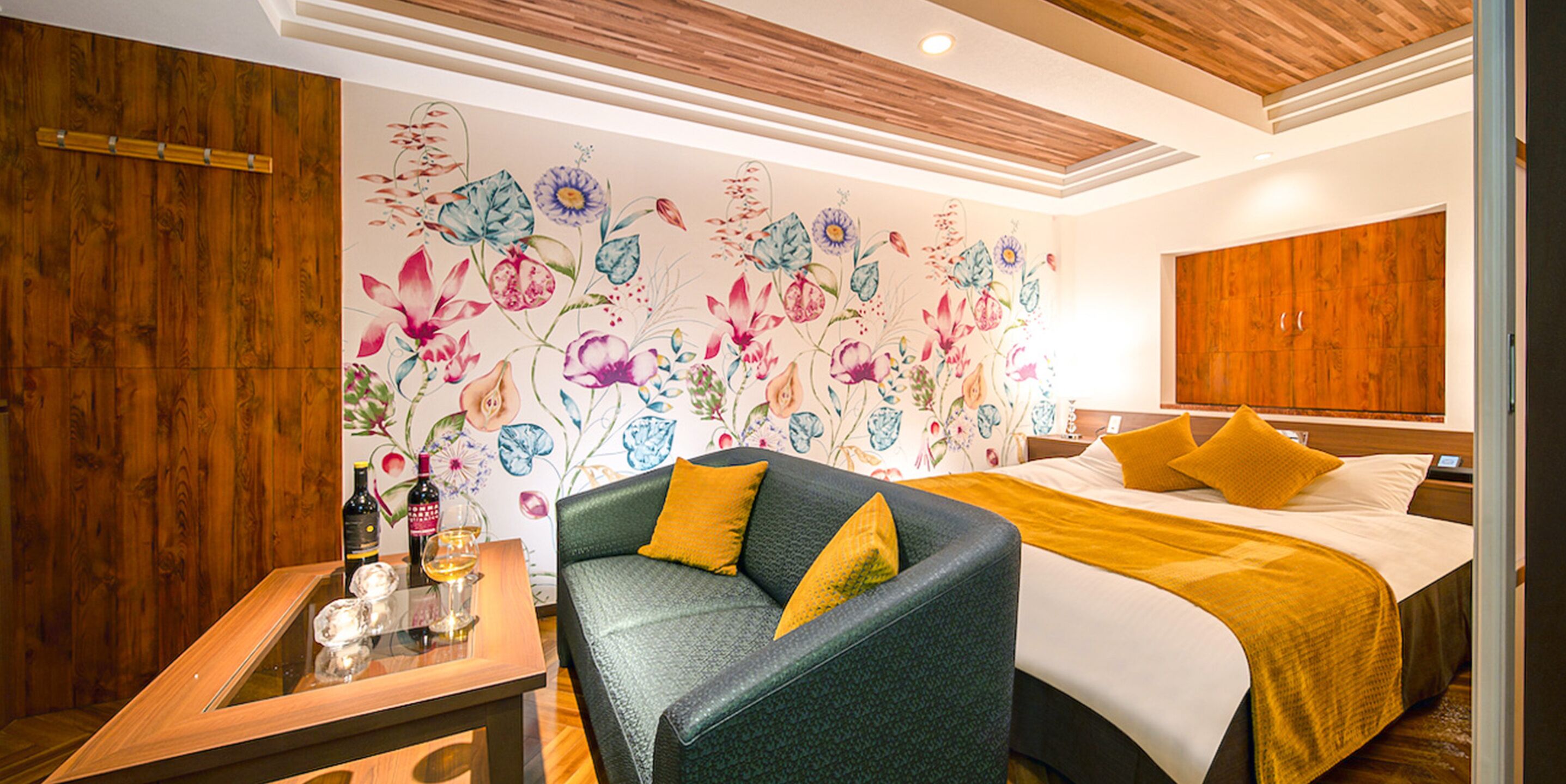 A hotel room with a floral wallpaper, a green sofa with yellow cushions, a wooden side table with a wine bottle and glass, and a bed with yellow bedding.
