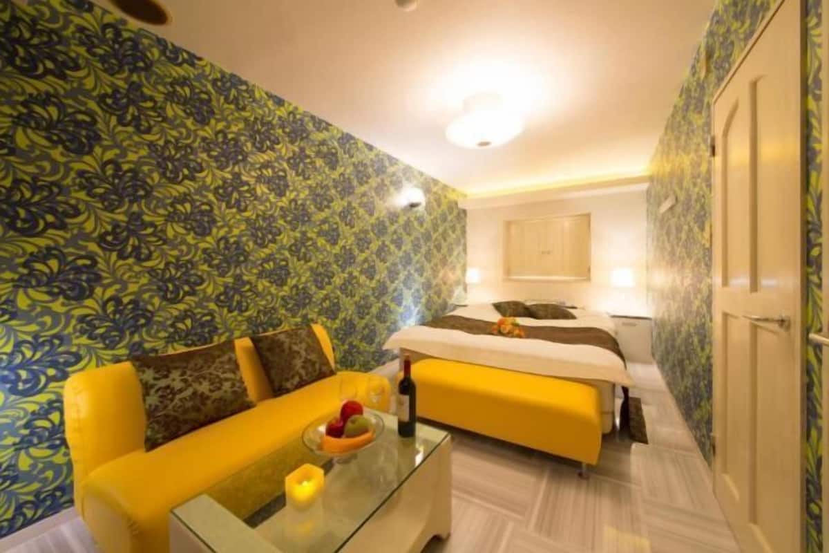 A hotel room with a patterned wallpaper, a yellow sofa, a glass coffee table with fruit and a bottle, and a bed with a white headboard.
