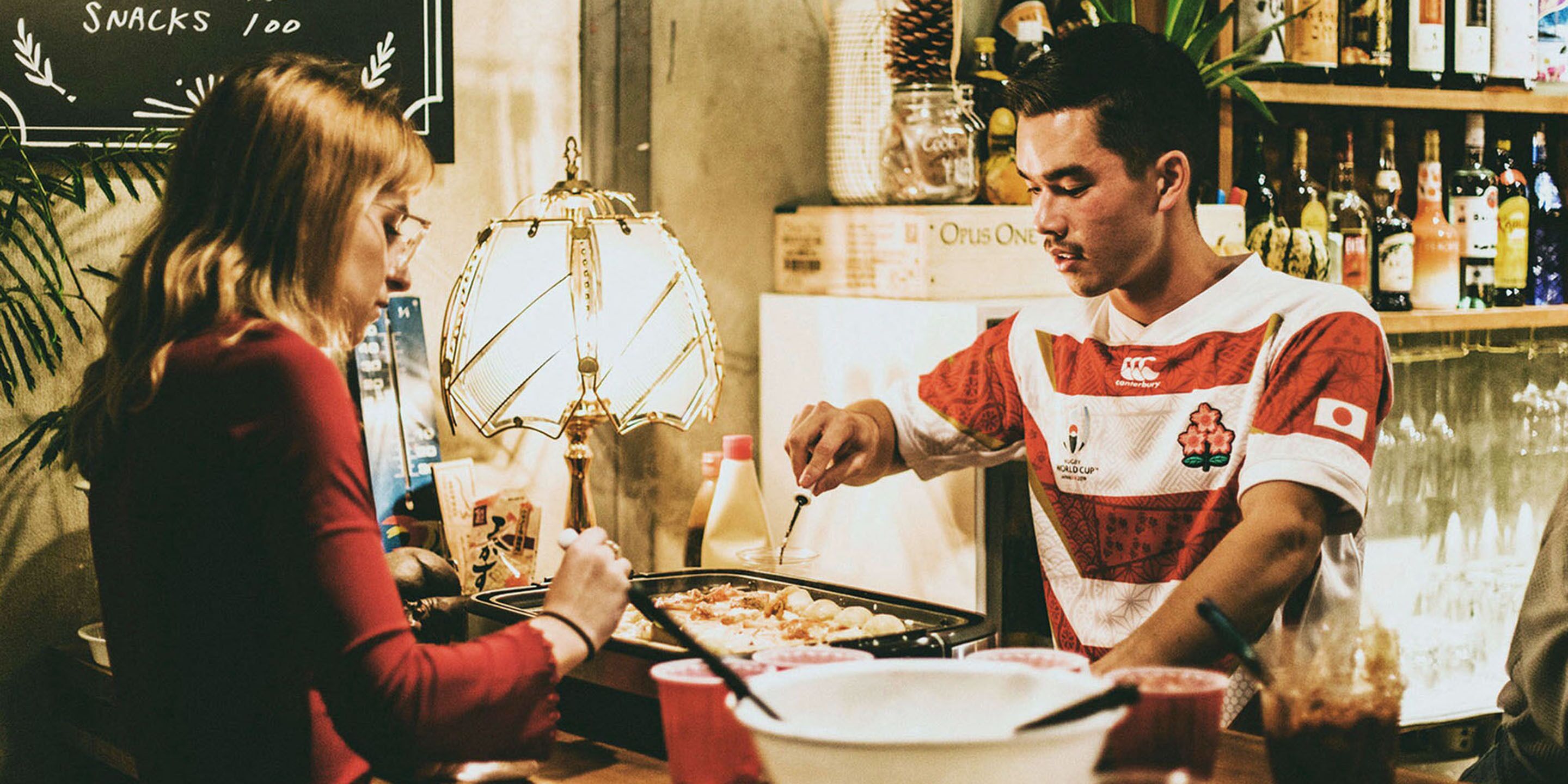 Una persona cocina en la encimera de la cocina, con una sartén, una botella de salsa y una taza roja sobre la mesa.