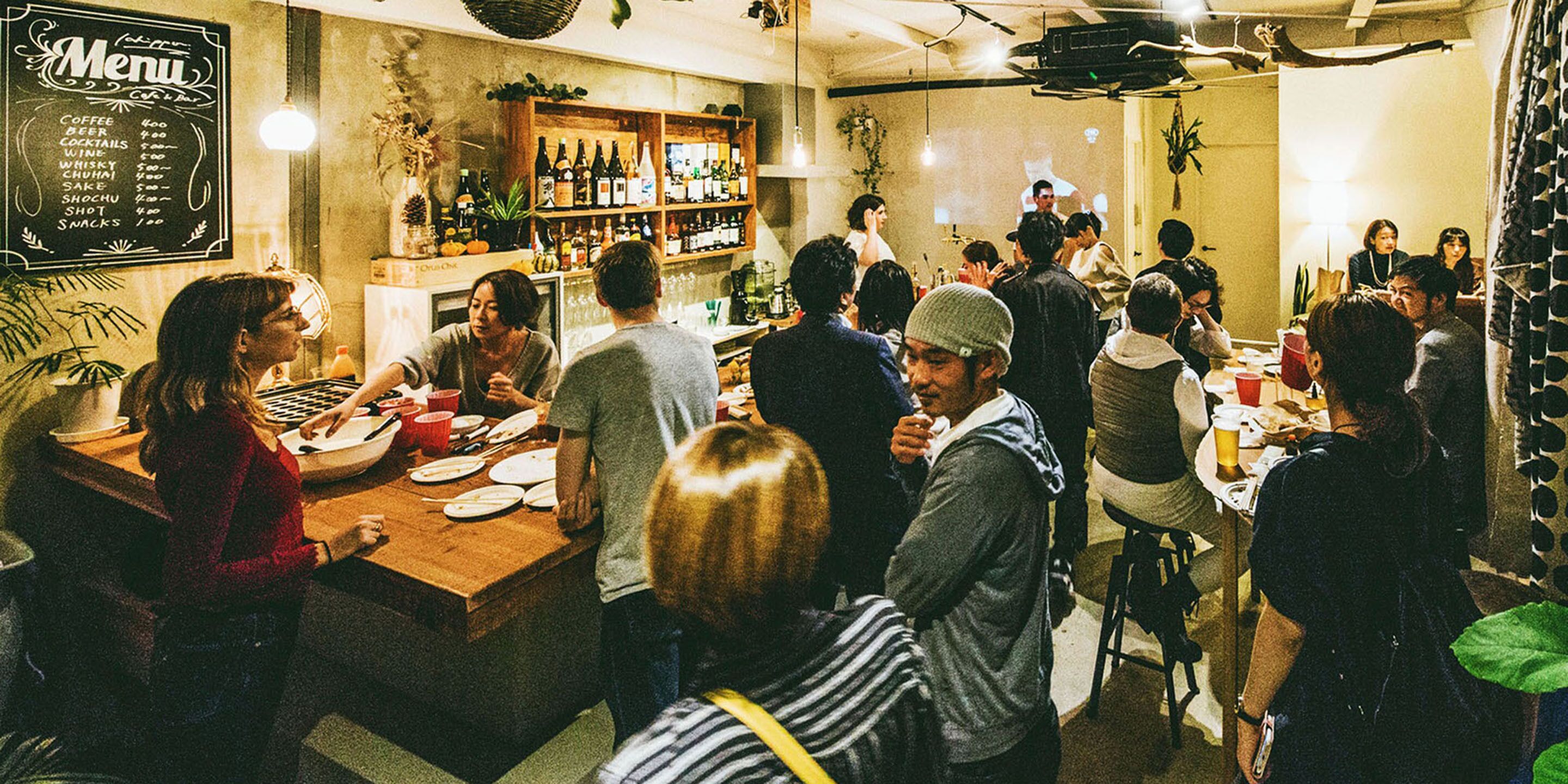 Un bar bondé, avec un menu affiché au mur, des étagères remplies de bouteilles, et des clients qui mangent et discutent.